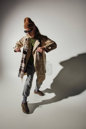 Alter mit Stilkonzept, durchtrainierter Senior-Hipster mit dunkler Sonnenbrille, Beanie-Hut, beigem Trenchcoat und kariertem Schal in stilvoller Pose auf grauem Hintergrund mit Schatten