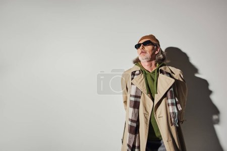 älterer Herr in stylischer Freizeitkleidung und dunkler Sonnenbrille, der vor grauem Hintergrund mit Schatten steht und wegschaut, Hipster-Trend, Beanie-Hut, beiger Trenchcoat, Mode- und Alterskonzept