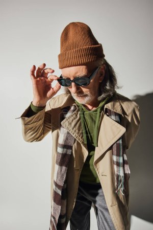 Photo for Bearded and senior man in beanie hat, plaid scarf and beige trench coat adjusting dark sunglasses on grey background with shadow, hipster fashion, stylish aging concept - Royalty Free Image