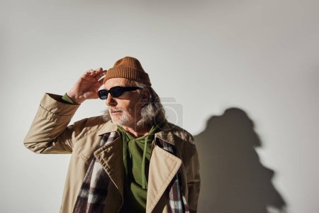 anciano hombre mayor de pelo gris y barbudo en gafas de sol oscuras, gabardina beige y bufanda a cuadros tocando sombrero de gorro y mirando hacia otro lado sobre fondo gris con sombra, estilo hipster, individualidad