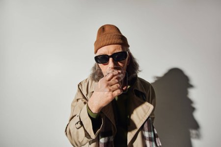trendy senior man in casual hipster style clothes holding hand near face and looking at camera on grey background with shadow, dark sunglasses, beanie hat, beige trench coat, fashion and age concept