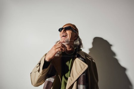 glücklich und stilvoll alterndes Konzept, aufgeregter Senior im Hipster-Stil mit dunkler Sonnenbrille, Beanie-Hut und beigem Trenchcoat schaut weg und lacht auf grauem Hintergrund mit Schatten