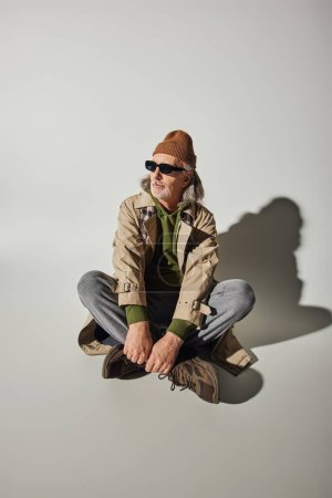 Téléchargez les photos : Longueur totale de l'homme âgé en costume de style hipster et lunettes de soleil sombres assis avec jambes croisées sur fond gris avec ombre, bonnet chapeau, trench coat beige, baskets, concept de mode et d'âge - en image libre de droit