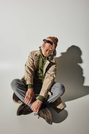 homme hipster senior en bonnet, lunettes de soleil sombres et trench coat beige assis avec jambes croisées et regardant loin sur fond gris avec ombre, concept de vieillissement à la mode