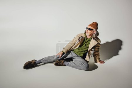 full length of fashionable senior male model sitting and looking away on grey background with shadow, dark sunglasses, beanie hat, beige trench coat, hipster style, positive aging concept