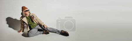 Photo for Full length of elderly man in dark sunglasses, beanie hat, beige trench coat and sneakers sitting and looking away on grey background with shadow, hipster fashion, banner with copy space - Royalty Free Image