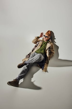 top view of trendy hipster man in beanie hat, beige trench coat and sneakers adjusting dark sunglasses and looking away while laying on grey background with shadow, stylish aging concept