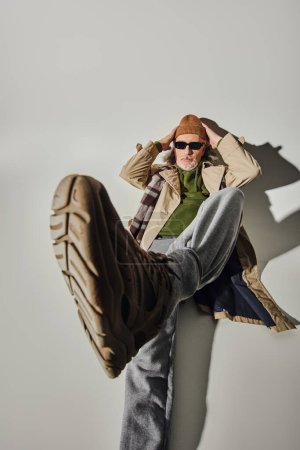 Photo for Full length of cool and bearded senior man in dark sunglasses and beanie hat laying with outstretched leg on grey background with shadow, hipster style, individuality, top view - Royalty Free Image