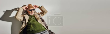 hombre de estilo hipster de moda en gafas de sol oscuras y gabardina beige ajustando sombrero de gorro mientras posa sobre fondo gris con sombra, envejecimiento con concepto de estilo, bandera