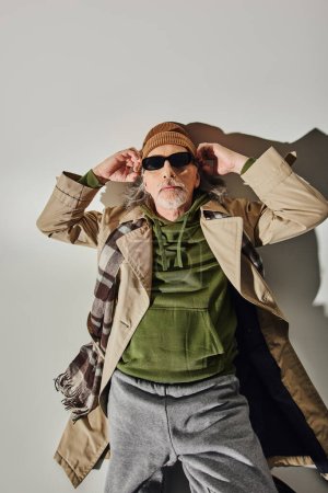 Draufsicht auf trendiges Senior-Model mit dunkler Sonnenbrille auf grauem Hintergrund mit Schatten, gealterter Hipster-Mann, Beanie-Hut, grüner Kapuzenpulli, beiger Trenchcoat, Alterung mit Stilkonzept