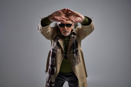 Téléchargez les photos : Tournage de mode de l'homme hipster senior en vêtements décontractés à la mode et lunettes de soleil sombres debout avec les mains tendues serrées et en regardant la caméra sur fond gris, concept de mode et d'âge - en image libre de droit