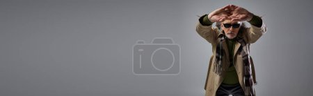 trendiger Mann im Hipster-Stil mit dunkler Sonnenbrille und beigem Trenchcoat, der mit geballten ausgestreckten Händen auf grauem Hintergrund posiert, stilvolles Altern, Modeshooting, Banner mit Kopierraum