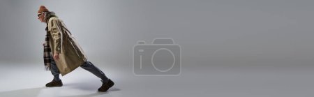Foto de Vista lateral del hombre anciano en traje de estilo hipster de moda pisando fondo gris, gafas de sol oscuras, gorro sombrero, gabardina beige, zapatillas de deporte, envejecimiento con el concepto de estilo, pancarta con espacio de copia - Imagen libre de derechos