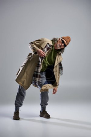 Photo for Full length of expressive senior man in dark sunglasses, beanie hat, beige trench coat and plaid scarf looking at camera on grey background, hipster fashion, stylish pose, trendy aging concept - Royalty Free Image