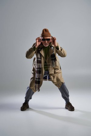 Photo for Full length of fashionable senior model in stylish pose, aged hipster man in beanie hat and beige trench coat adjusting dark sunglasses and looking at camera on grey background - Royalty Free Image