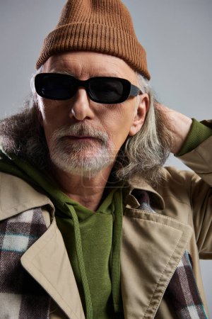 Ausdrucksstarke Persönlichkeit, Porträt eines älteren Mannes im Hipster-Stil mit Mütze, dunkler Sonnenbrille, beigem Trenchcoat und kariertem Schal vor der Kamera auf grauem Hintergrund, trendiger Lebensstil 