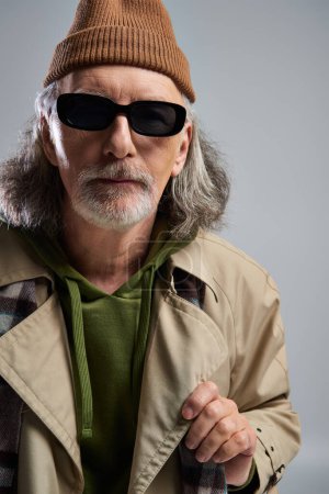 Porträt eines grauhaarigen und bärtigen Mannes mit Hut, dunkler Sonnenbrille und beigem Trenchcoat, der auf grauem Hintergrund in die Kamera blickt, Modell im Hipster-Stil, modisches Alterskonzept
