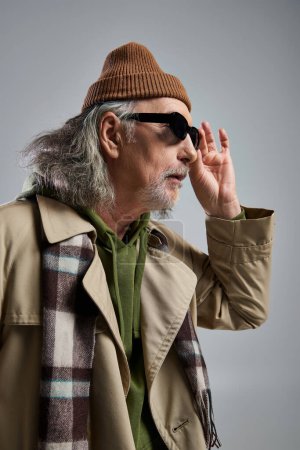 Téléchargez les photos : Portrait d'homme âgé étonné en bonnet, trench coat beige et foulard à carreaux ajustant les lunettes de soleil sombres et regardant loin sur fond gris, mode hipster, concept de vieillissement élégant et positif - en image libre de droit