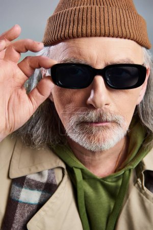Porträt eines älteren Mannes mit gepflegtem Bart, mit Mütze und beigem Trenchcoat, dunkler Sonnenbrille und Blick in die Kamera auf grauem Hintergrund, Hipster-Mode, Individualität, Altern mit Stil