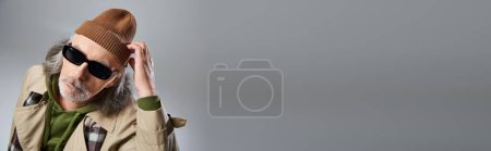 Téléchargez les photos : Portrait de mannequin homme senior en lunettes de soleil sombres et trench coat beige ajustant les lunettes de soleil sombres et regardant la caméra sur fond gris, homme hipster âgé, bannière avec espace de copie - en image libre de droit