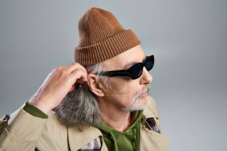 portrait of elderly and bearded hipster style senior man in dark sunglasses, beanie hat and beige trench coat adjusting grey hair and looking away on grey background, fashionable aging concept
