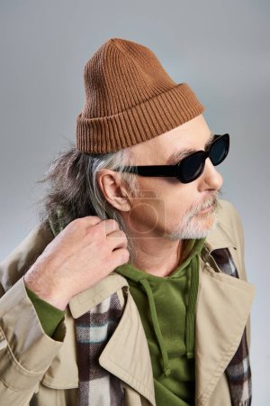 portrait of aged hipster man in dark sunglasses, beanie hat, green hoodie and beige trench coat looking away while standing on grey background, aging with style concept, fashion shoot