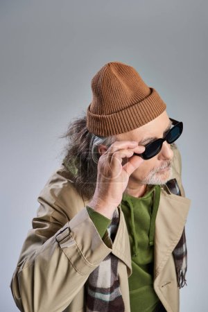 high angle view of fashionable and bearded aged man in beanie hat and beige coat adjusting dark sunglasses and looking away on grey background, trendy lifestyle and aging concept
