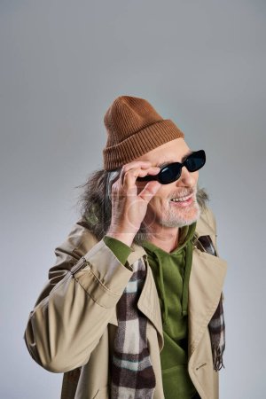 Porträt eines älteren und fröhlichen bärtigen Mannes im Hipster-Stil mit Hut und beigem Trenchcoat, der eine dunkle Sonnenbrille trägt, lacht und vor grauem Hintergrund wegschaut