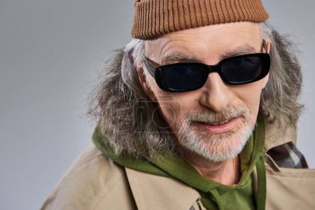 portret uśmiechniętego starszego mężczyzny z siwymi włosami i zadbaną brodą, w ciemnych okularach przeciwsłonecznych, czapce z kapelusza i płaszczu z rowu patrzącego na kamerę na szarym tle, moda hipsterska, szczęśliwa i modna koncepcja starzenia się
