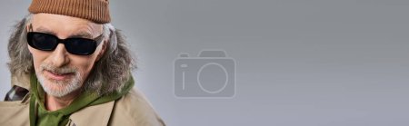 Ausdrucksstarke Individualität, Porträt eines lächelnden Seniors mit Mütze, dunkler stylischer Sonnenbrille und beigem Trenchcoat, der auf grauem Hintergrund in die Kamera lächelt, positives Altern, Banner mit Kopierraum