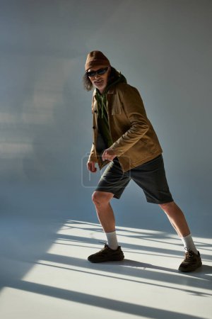 voller Länge fröhlicher älterer Herr im Hipster-Outfit posiert auf grauem Hintergrund mit Beleuchtung und Blick in die Kamera, dunkle Sonnenbrille, Hut, Jacke und Shorts, Modeshooting