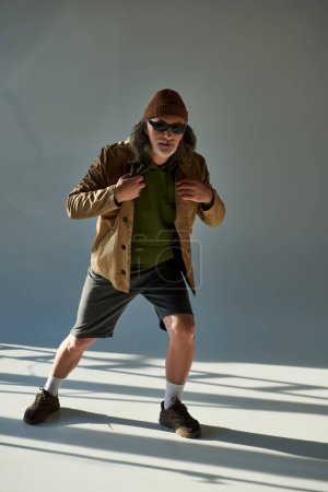 full length of elderly and hipster style man in beanie hat, dark sunglasses, jacket and shorts looking at camera on grey background with lighting, stylish pose, trendy aging concept