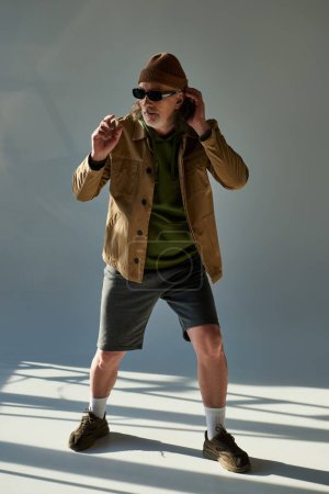 fashion shoot of elderly man in dark sunglasses, beanie hat, jacket and shorts looking away on grey background with lighting, hipster trend, fashionable aging concept, full length view