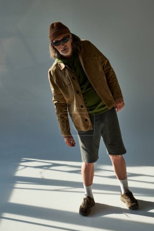 pełna długość męskiego modelu patrzącego w kamerę na szarym tle z oświetleniem, starszego hipstera w ciemnych okularach przeciwsłonecznych, czapce z daszkiem, kurtce i spodenkach, modna koncepcja stylu życia
