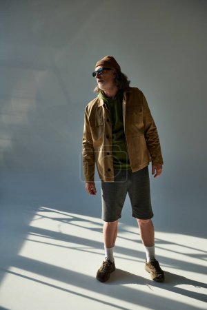 vollbärtiger älterer Mann mit dunkler Sonnenbrille, Hut, Jacke und kurzen Hosen auf grauem Hintergrund mit Beleuchtung, Hipster-Trend, ausdrucksstarke Persönlichkeit