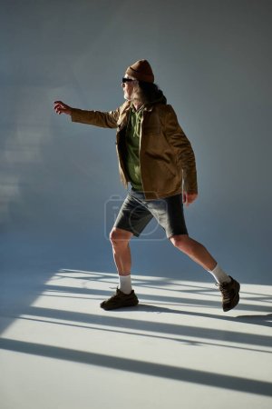 full length of senior man in fashionable casual clothes, dark sunglasses, beanie hat, jacket and shorts pointing with hand and looking away on grey background with lighting, aging with style concept