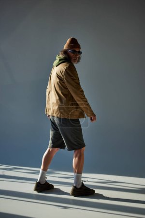 Älterer Herr im Hipster-Look mit Hut, dunkler Sonnenbrille, Jacke und kurzen Hosen, der wegschaut, während er auf grauem Hintergrund mit Beleuchtung steht, modisches Alterskonzept