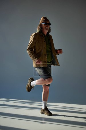 volle Länge von lächelnden und stilvollen männlichen Senior-Modell posiert auf einem Bein auf grauem Hintergrund mit Beleuchtung, Hipster-Mode, dunkle Sonnenbrille, Jacke und Shorts, glücklich und modisch alternden Konzept