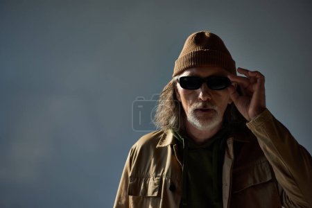 modisch und hipster gestylt Senior mit Hut und brauner Jacke, dunkle Sonnenbrille und Blick in die Kamera auf grauem Hintergrund, alterndes Lebensstil-Konzept