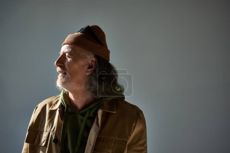grauhaariger, bärtiger und zufriedener älterer Mann mit Hut und brauner Jacke, der mit geschlossenen Augen vor grauem Hintergrund steht, Hipster-Stil, fröhliches und modisches Alterskonzept