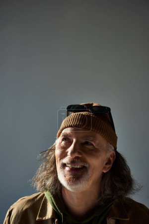 Photo for Portrait of cheerful and carefree senior man with grey hair and beard, wearing dark sunglasses on beanie hat, looking up on grey background, with copy space, happy and fashionable aging concept - Royalty Free Image