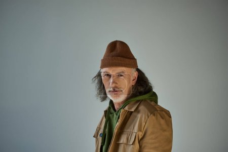grey haired, bearded and positive senior man in beanie hat and brown jacket looking at camera when standing on grey background, hipster style model, aging population lifestyle concept