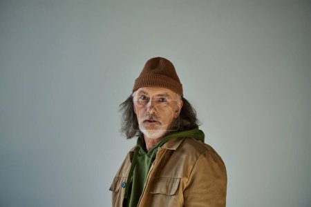 thoughtful and discouraged hipster style senior man in beanie hat and brown jacket looking at camera while standing on grey background, aging population lifestyle concept