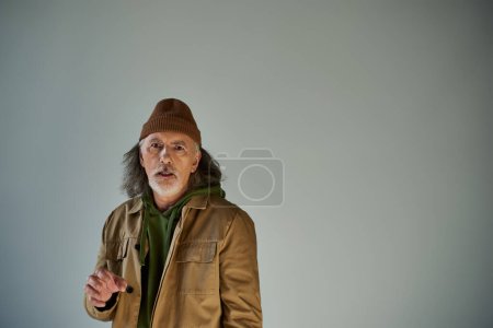 frustrated and worried elderly man with grey hair and beard, in beanie hat and brown jacket looking at camera on grey background, hipster style, aging population lifestyle concept
