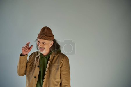 Optimistischer Senior-Hipster gestikulierend mit händennahem Kopf und lächelnd auf grauem Hintergrund mit Kopierraum, Beanie-Hut, brauner und trendiger Jacke, fröhliches und modisches Alterskonzept