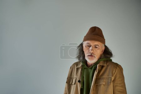 molesto hombre mayor de pelo gris y barbudo en gorro sombrero y chaqueta marrón de pie sobre fondo gris, estilo hipster, personalidad expresiva, envejecimiento concepto de estilo de vida de la población