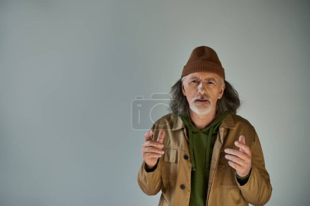 Aufgebrachter und besorgter älterer bärtiger Mann mit Mütze und brauner Jacke gestikuliert und blickt in die Kamera auf grauem Hintergrund, Hipster-Klamotten, alterndes Lebensstil-Konzept