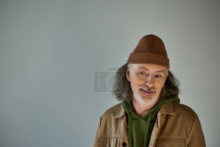 grauhaariger und bärtiger älterer Herr, nachdenklich und lächelnd, mit Hut und brauner Jacke auf grauem Hintergrund, Hipster-Mode, fröhliches und modisches Alterskonzept