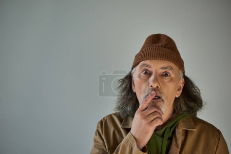 impressed and shocked senior man with grey hair and beard touching lips and looking at camera on grey background, beanie hat, brown jacket, hipster fashion, aging population lifestyle concept
