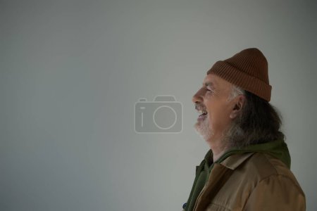 Téléchargez les photos : Vue latérale de l'homme âgé excité, aux cheveux gris et barbu en bonnet chapeau et veste brune riant sur fond gris, concept de vieillissement heureux et à la mode, espace de copie - en image libre de droit